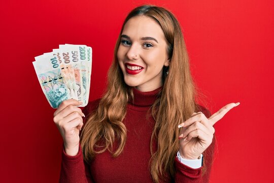 Young blonde woman holding czech koruna banknotes smiling happy pointing with hand and finger to the side