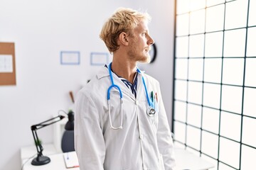 Young blond man wearing doctor uniform and stethoscope at clinic looking to side, relax profile pose with natural face and confident smile.