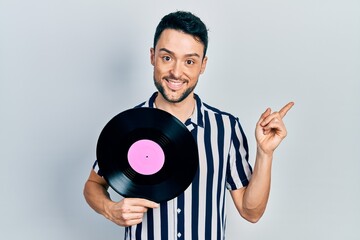 Young hispanic man holding vinyl disc smiling happy pointing with hand and finger to the side