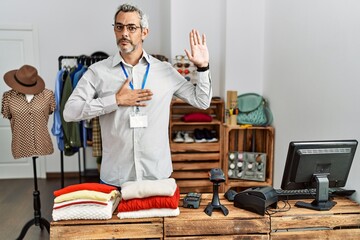 Middle age hispanic man working as manager at retail boutique swearing with hand on chest and open...