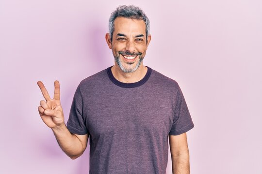 Handsome middle age man with grey hair wearing casual t shirt smiling with happy face winking at the camera doing victory sign. number two.
