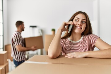 Young hispanic woman talking on the smartphone leaning on cardboard at new home.
