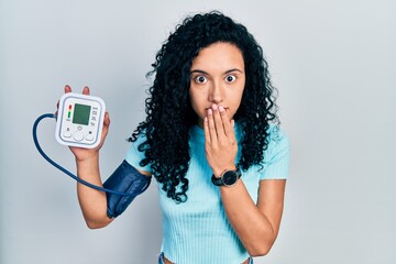 Young hispanic woman with curly hair using blood pressure monitor covering mouth with hand, shocked...