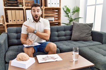 Handsome hispanic man working with rorschach test at psychology clinic surprised pointing with finger to the side, open mouth amazed expression.