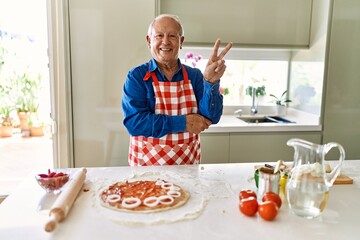 Senior man with grey hair cooking pizza at home kitchen smiling with happy face winking at the camera doing victory sign. number two.