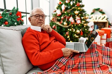 Senior man with grey hair using laptop sitting by christmas tree serious face thinking about question with hand on chin, thoughtful about confusing idea