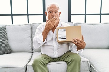 Senior man holding delivery package at home covering mouth with hand, shocked and afraid for mistake. surprised expression