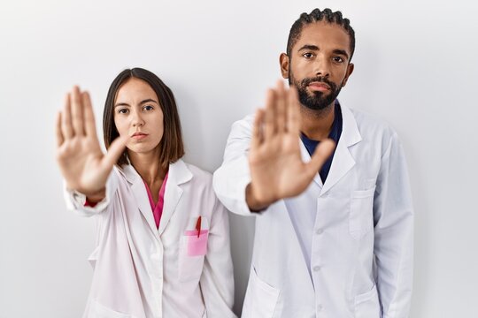 Young hispanic doctors standing over white background doing stop sing with palm of the hand. warning expression with negative and serious gesture on the face.