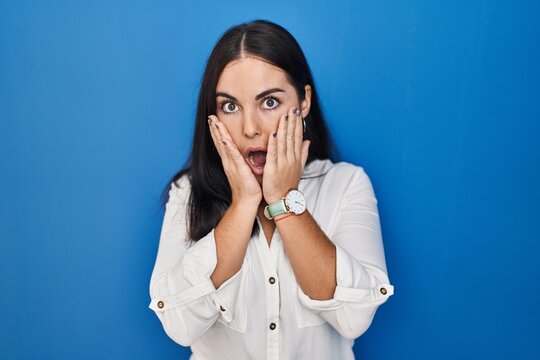 Young hispanic woman standing over blue background afraid and shocked, surprise and amazed expression with hands on face