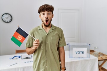 Young arab man at political campaign election holding afghanistan flag scared and amazed with open mouth for surprise, disbelief face