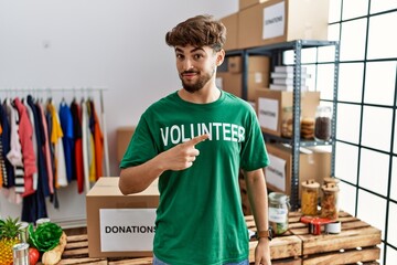 Young arab man wearing volunteer t shirt at donations stand pointing with hand finger to the side showing advertisement, serious and calm face