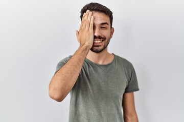Young hispanic man with beard wearing casual t shirt over white background covering one eye with hand, confident smile on face and surprise emotion.