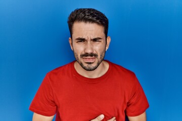 Fototapeta premium Young hispanic man with beard wearing red t shirt over blue background with hand on stomach because indigestion, painful illness feeling unwell. ache concept.
