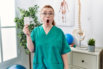 Young redhead physiotherapist woman holding prescription pills scared and amazed with open mouth for surprise, disbelief face