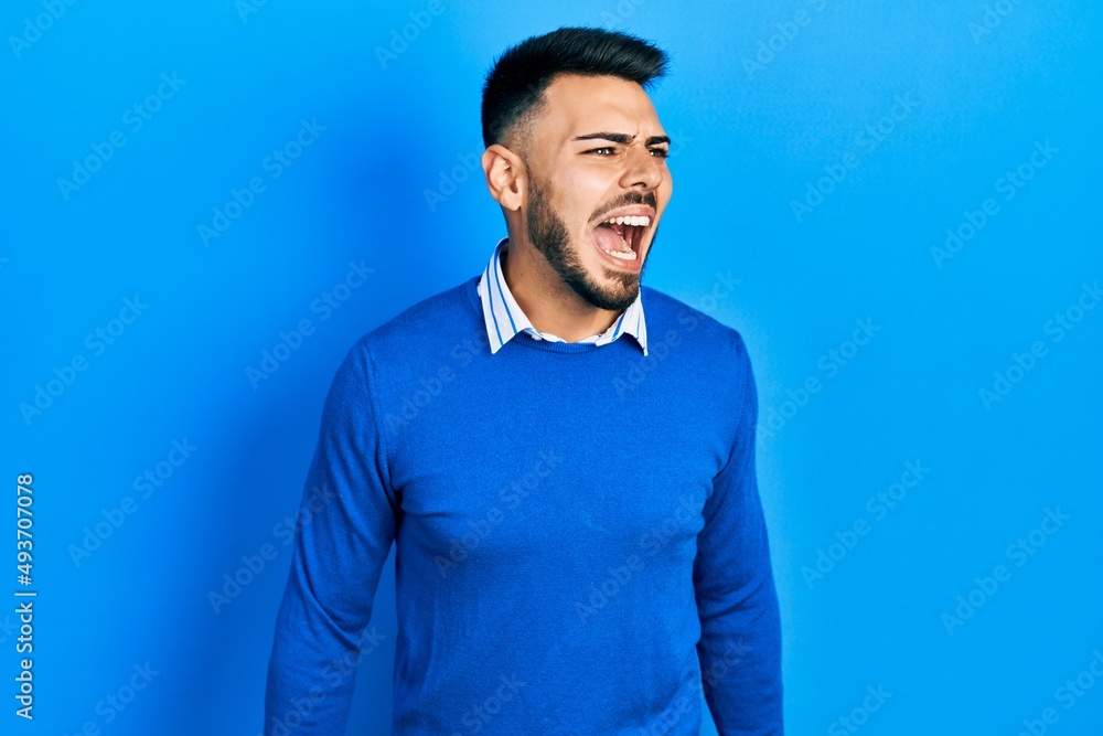 Wall mural Young hispanic man with beard wearing casual blue sweater angry and mad screaming frustrated and furious, shouting with anger. rage and aggressive concept.