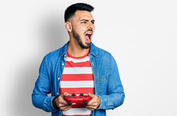 Young hispanic man with beard playing video game holding controller angry and mad screaming...