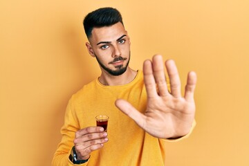 Young hispanic man with beard drinking whiskey shot with open hand doing stop sign with serious and...