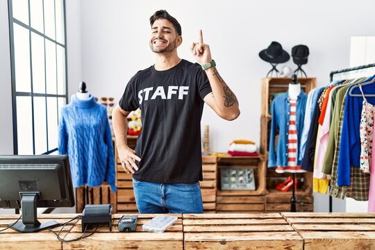 Young hispanic man working at retail boutique pointing finger up with successful idea. exited and happy. number one.