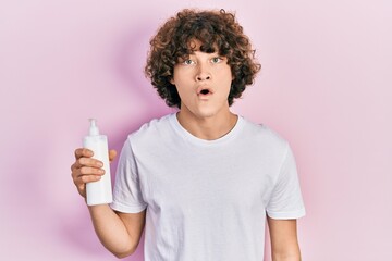 Handsome young man holding cosmetic moisturizer facial cream scared and amazed with open mouth for...
