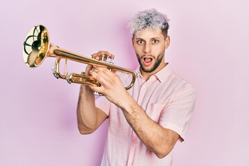 Young hispanic man with modern dyed hair playing trumpet afraid and shocked with surprise and...