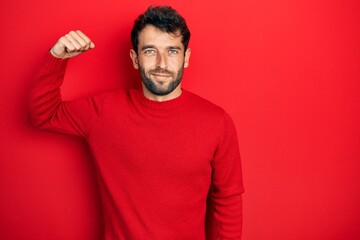 Handsome man with beard wearing casual red sweater strong person showing arm muscle, confident and proud of power