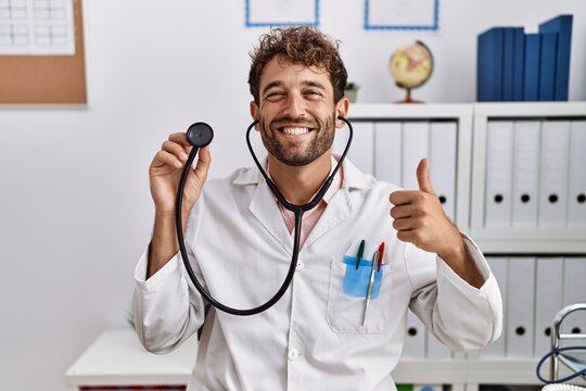 Young hispanic doctor man wearing doctor uniform holding stethoscope at clinic smiling happy and positive, thumb up doing excellent and approval sign