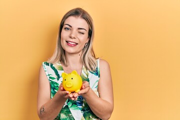 Beautiful caucasian woman holding piggy bank winking looking at the camera with sexy expression, cheerful and happy face.