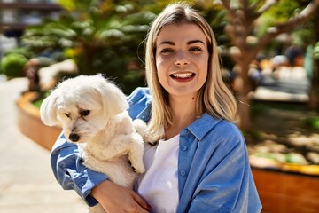 Young blonde girl smiling happy holding dog at the city.