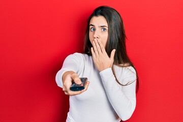 Naklejka premium Young brunette teenager holding television remote control covering mouth with hand, shocked and afraid for mistake. surprised expression