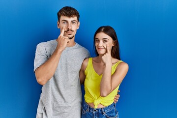 Young hispanic couple standing together over blue background pointing to the eye watching you gesture, suspicious expression