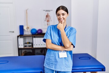 Young hispanic woman wearing physiotherapist uniform standing at clinic looking confident at the camera smiling with crossed arms and hand raised on chin. thinking positive.