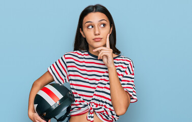 Young hispanic girl holding motorcycle helmet serious face thinking about question with hand on chin, thoughtful about confusing idea