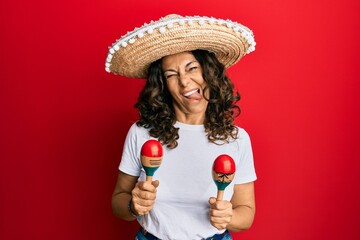 Middle age hispanic woman holding mexican hat playing maracas sticking tongue out happy with funny...