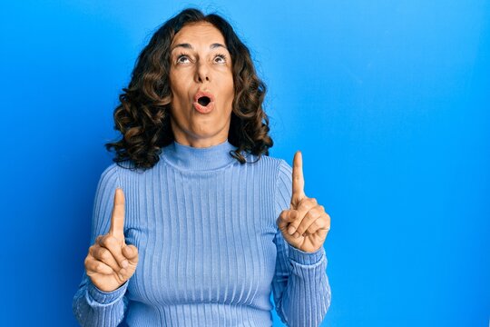 Middle age hispanic woman wearing casual clothes amazed and surprised looking up and pointing with fingers and raised arms.