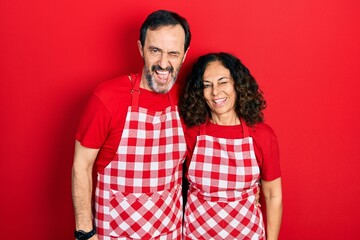 Middle age couple of hispanic woman and man wearing cook apron winking looking at the camera with sexy expression, cheerful and happy face.