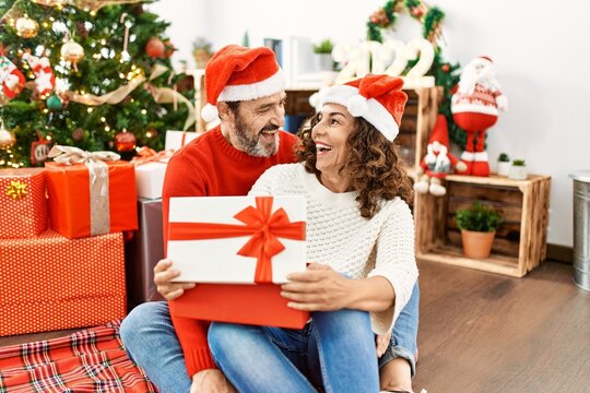 Middle age hispanic couple wearing christmas hat. Sitting on the floor opening gift at home.