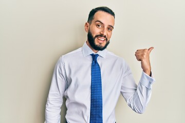 Young man with beard wearing business tie smiling with happy face looking and pointing to the side with thumb up.