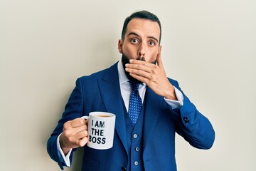 Young man with beard drinking from i am the boss coffee cup covering mouth with hand, shocked and afraid for mistake. surprised expression