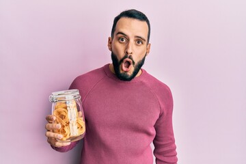 Young man with beard holding jar with pasta scared and amazed with open mouth for surprise,...