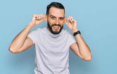 Young man with beard wearing casual white t shirt smiling pulling ears with fingers, funny gesture....