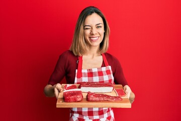 Young caucasian blonde woman holding board with raw meat winking looking at the camera with sexy...