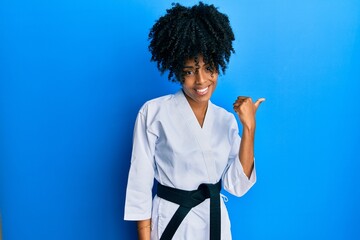 African american woman with afro hair wearing karate kimono and black belt smiling with happy face looking and pointing to the side with thumb up.