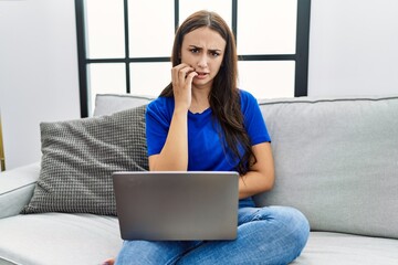Naklejka premium Young brunette woman using laptop at home looking stressed and nervous with hands on mouth biting nails. anxiety problem.