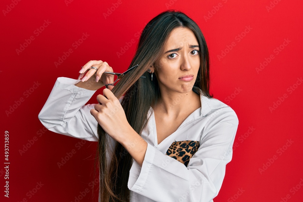 Sticker Young hispanic girl cutting hair using scissors depressed and worry for distress, crying angry and afraid. sad expression.