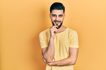 Handsome man with beard wearing casual yellow t shirt looking confident at the camera with smile with crossed arms and hand raised on chin. thinking positive.