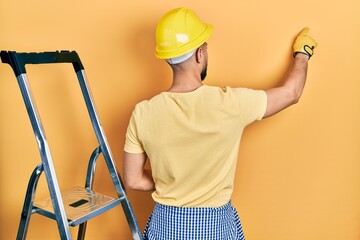 Handsome man with beard by construction stairs wearing hardhat posing backwards pointing ahead with finger hand