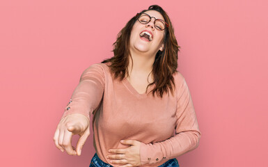 Young plus size woman wearing casual clothes and glasses laughing at you, pointing finger to the...