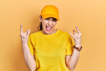 Young hispanic woman wearing delivery uniform and cap shouting with crazy expression doing rock symbol with hands up. music star. heavy concept.
