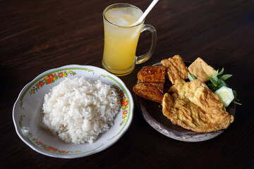Eggs, tempeh and eggplant penyet with white rice and orange juice on a wooden table, Indonesian cuisine