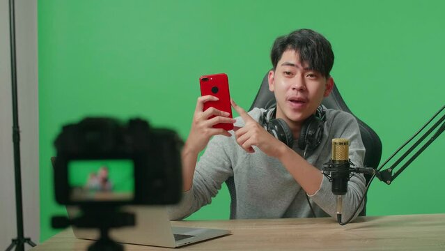 A Smiling Asian Man With Computer Reviewing Mobile Phone While Shooting Video By Camera On Green Screen Background
